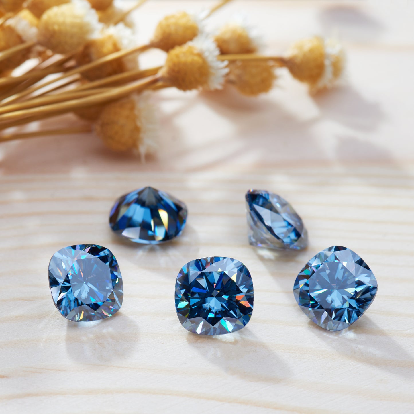 a collection of blue cushion cut moissanite stones on light wooden surface, close up shot, dried flowers as secondary motif