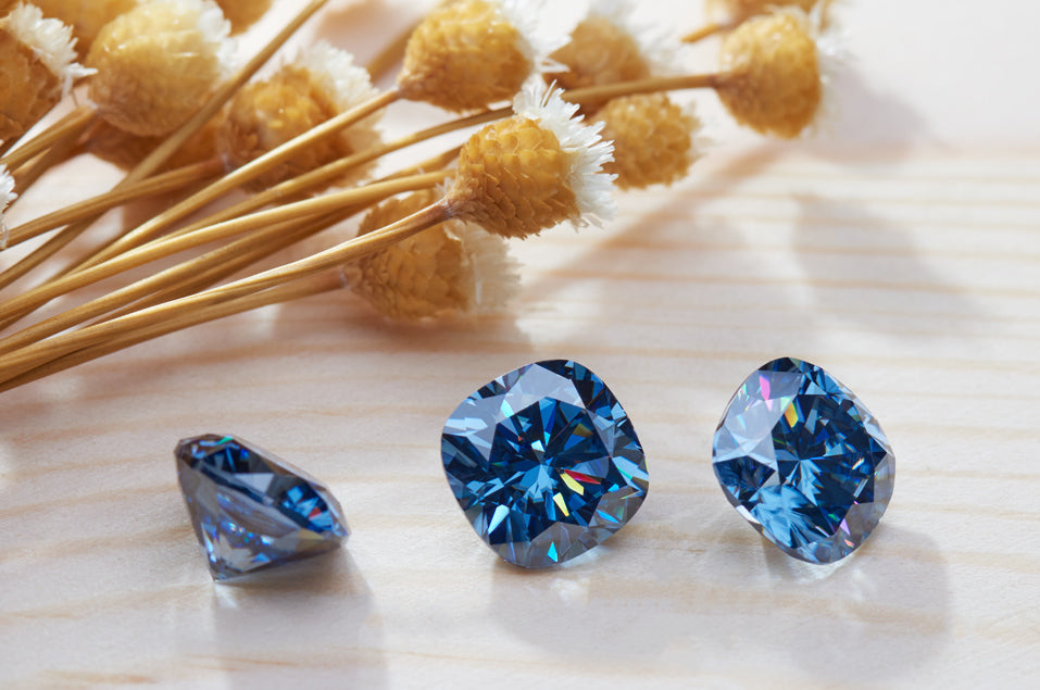 a trio of blue cushion cut moissanite stones on light wooden surface, close up shot, dried flowers as secondary motif