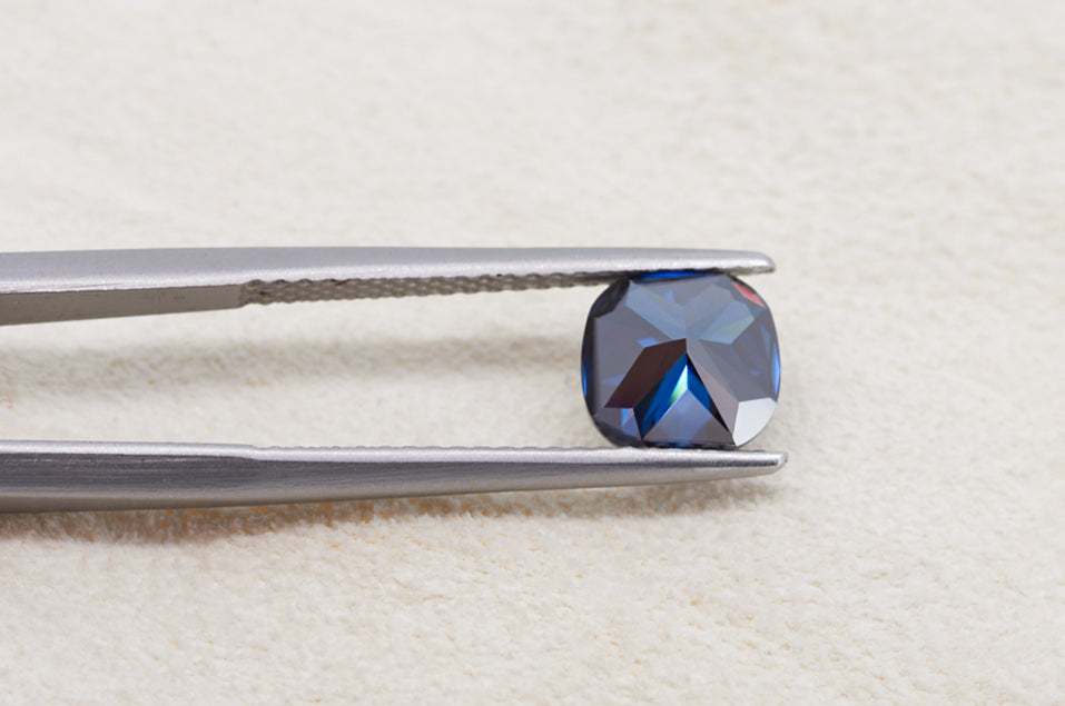 dark blue cushion cut moissanite stone between tweezers on beige fur background, back side