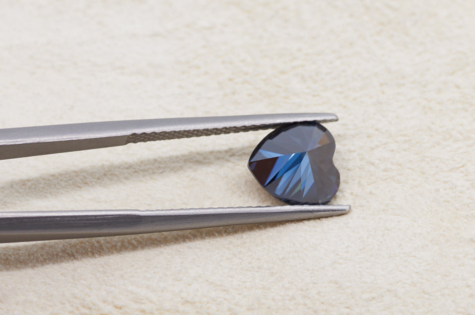 dark blue heart cut moissanite stone between tweezers on beige fur background, backside