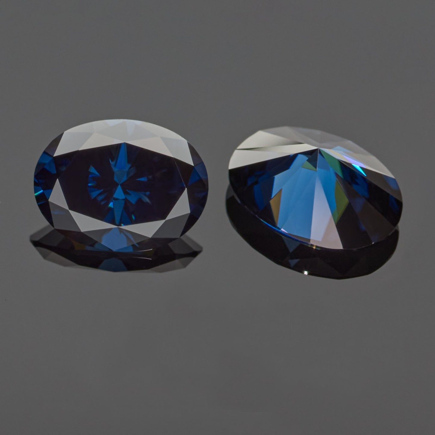 a pair of dark blue oval cut moissanite stones on reflective grey background, top and bottom view