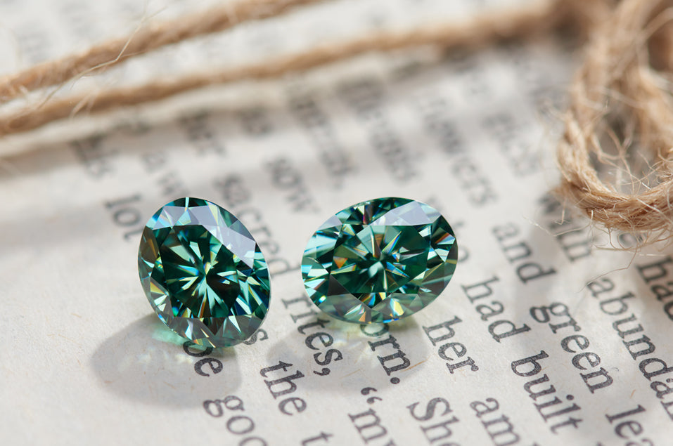 a pair of green oval cut moissanite stones, background is a book page and a piece of yarn