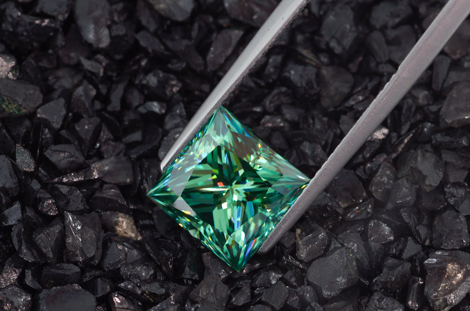 green princess cut moissanite stone between tweezers on black finely crushed rock