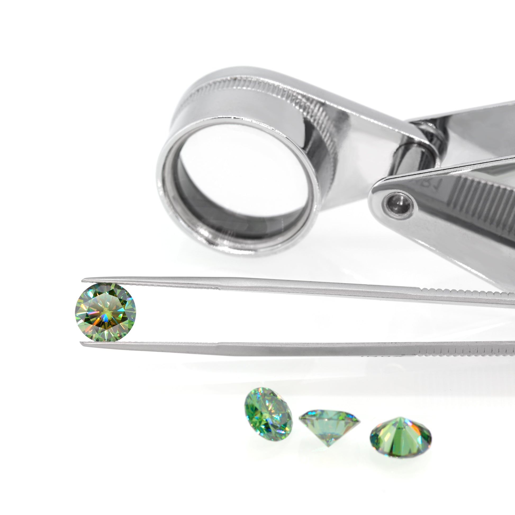 green round cut moissanite stone in tweezers, a few other green gemstones in foreground and a magnifying glass in background